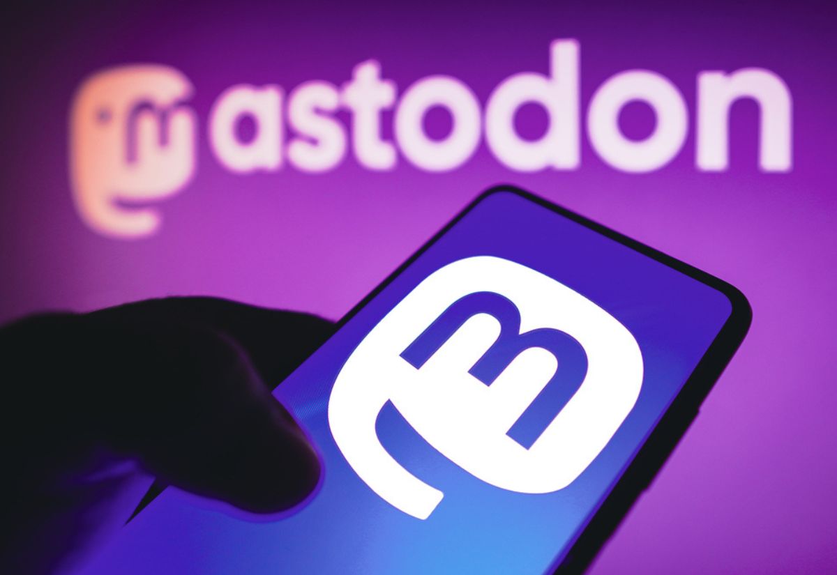 A smartphone screen displaying the Mastodon logo is held up against a blurred purple background with a larger Mastodon logo and text.