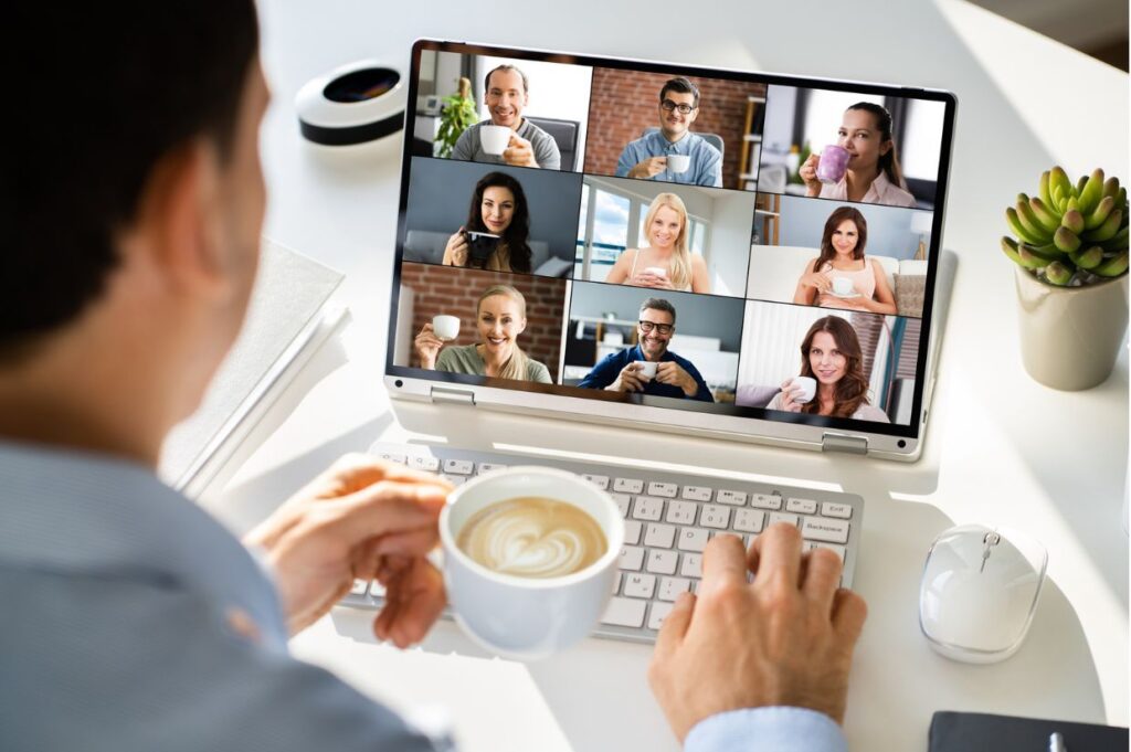 A person attends a virtual meeting on a laptop, engaging with multiple participants on screen while holding a cup of coffee.