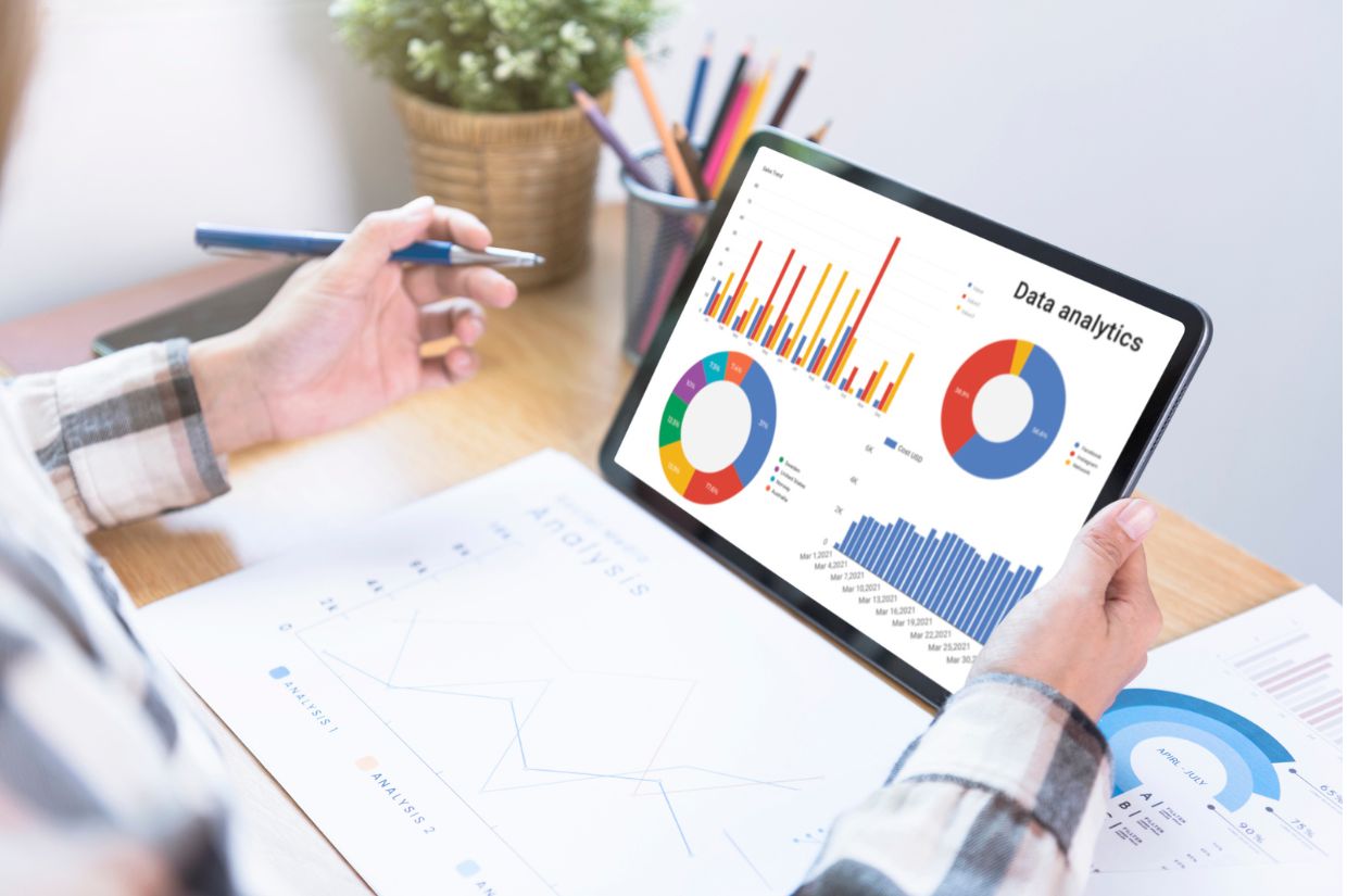 A person analyzes data on a tablet, featuring colorful graphs and charts, while taking notes at a desk with stationery and a small plant.