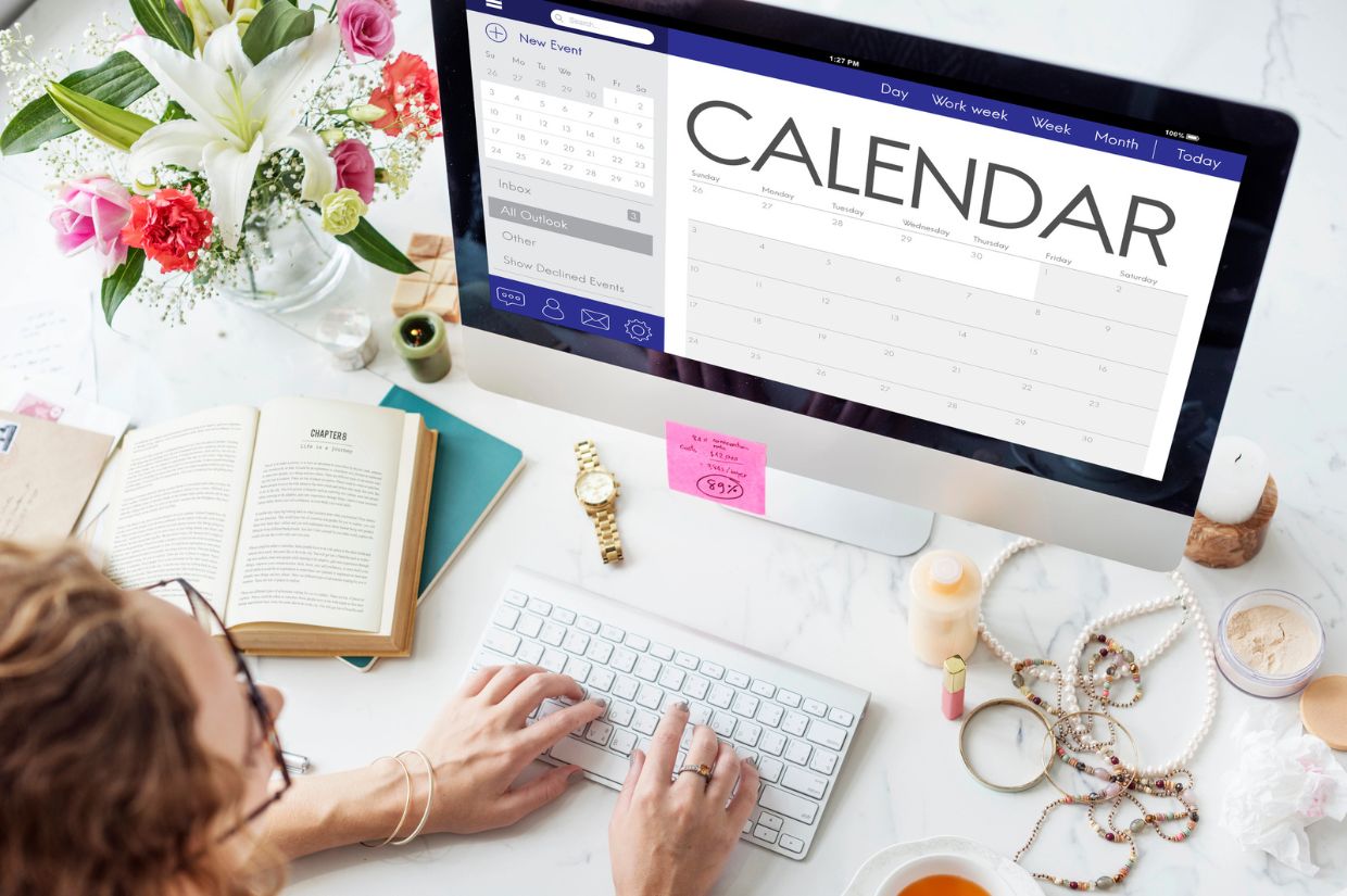 A person using a computer to view a calendar, surrounded by flowers, books, and jewelry on a desk.