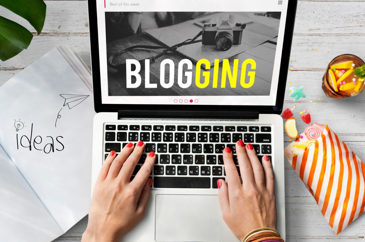 Hands typing on a laptop displaying "BLOGGING," next to a notepad with ideas and colorful snacks.