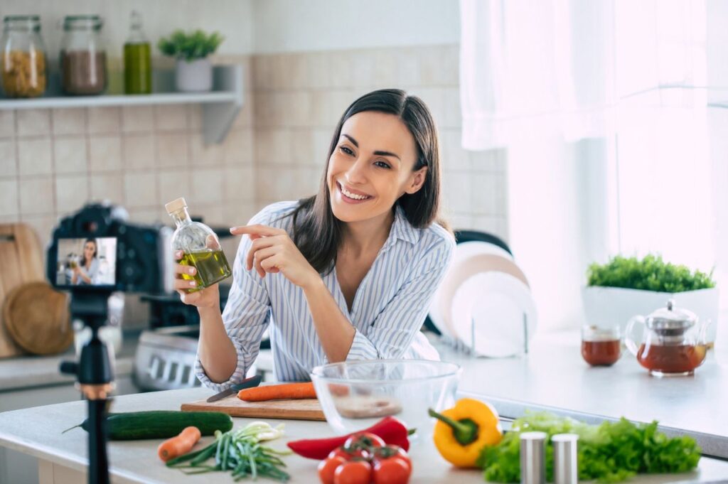 Professional beautiful happy young woman is blogging for her kitchen channel about healthy living in the kitchen of her home and looking on camera on a tripod.