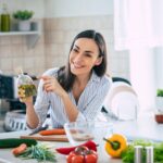 Professional beautiful happy young woman is blogging for her kitchen channel about healthy living in the kitchen of her home and looking on camera on a tripod.