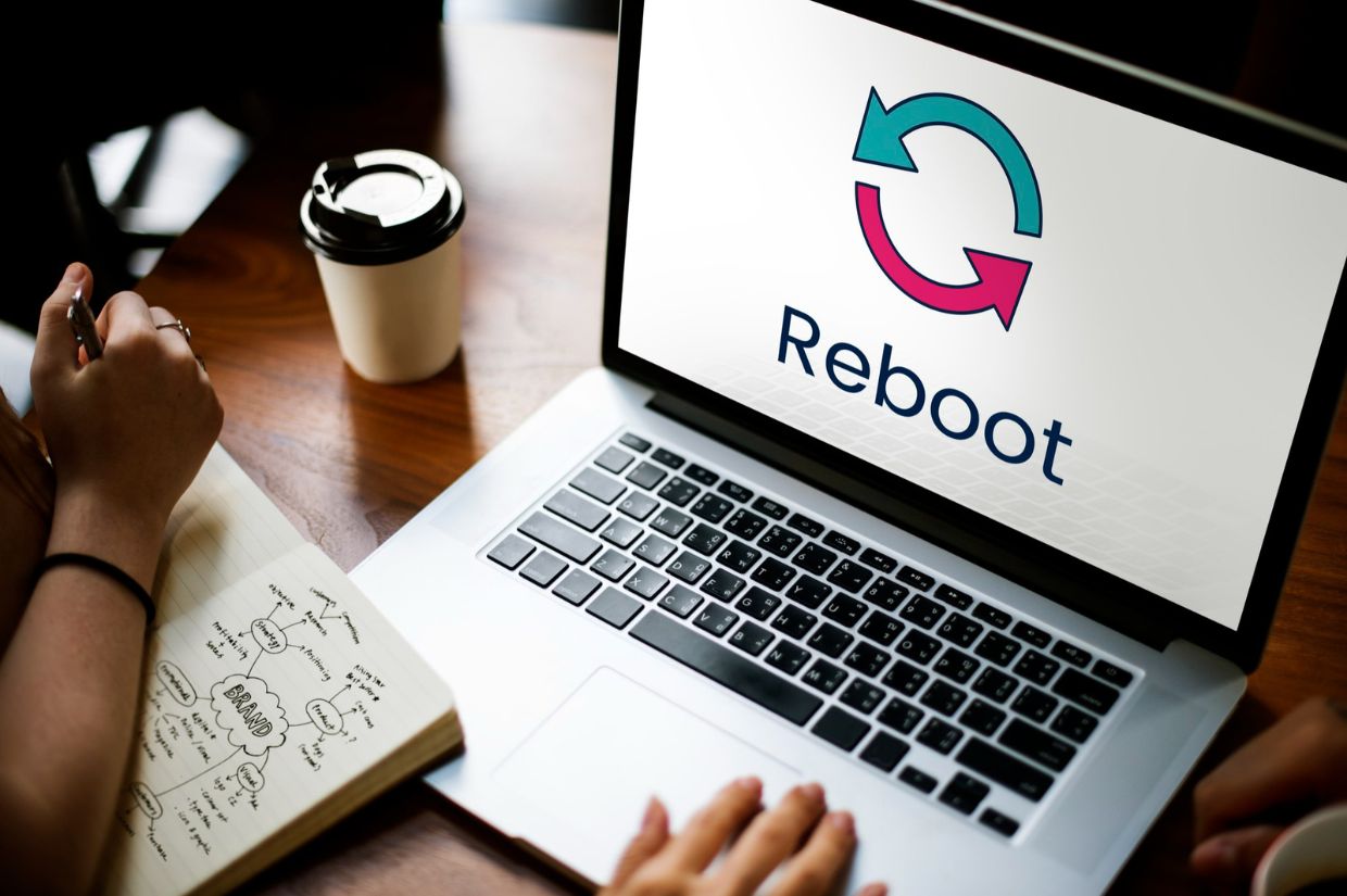 A laptop displaying the "Reboot" logo on a wooden table, accompanied by a coffee cup and a notebook with notes.