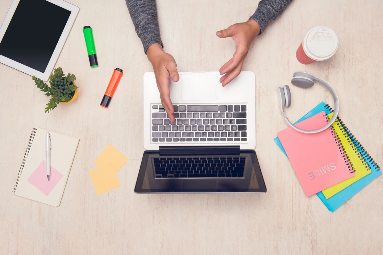 A top view of a workspace with a laptop, notebooks, stationery, a coffee cup, and a person gesturing with their hands.