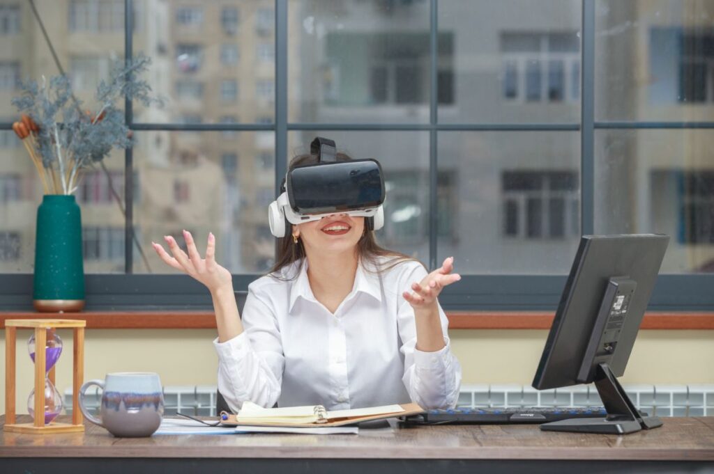 A young woman in a white shirt sits at a desk, smiling while wearing virtual reality (VR) glasses.