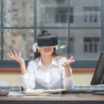 A young woman in a white shirt sits at a desk, smiling while wearing virtual reality (VR) glasses.