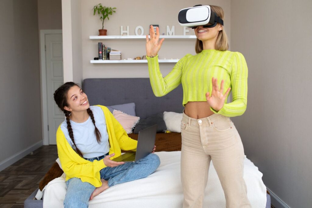 Two young women are in a bedroom. One woman is sitting on a bed with a laptop, smiling and looking at the other. The second woman, standing and wearing a VR headset, is engaged in a virtual experience, raising her hands as if interacting with something. 