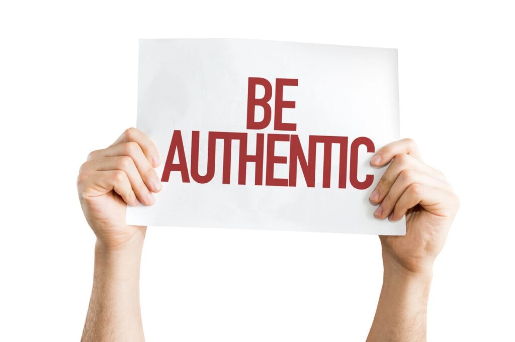 Two hands holding a white sign with the words 'BE AUTHENTIC' written in bold red capital letters.