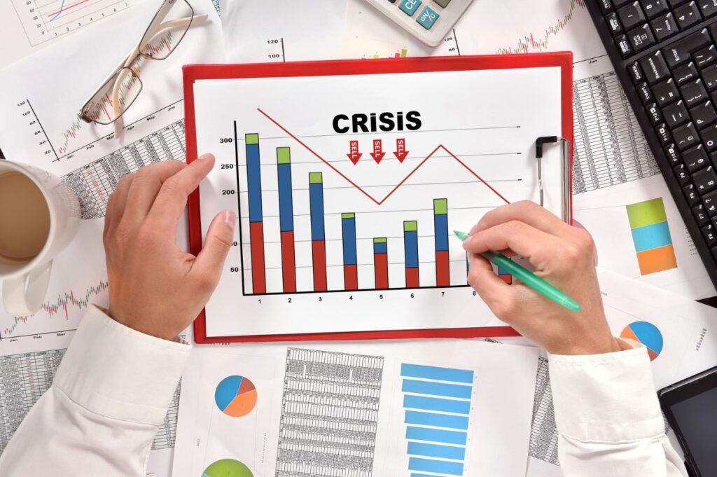 Person analyzing a bar and line chart labeled 'Crisis' on a clipboard, surrounded by financial documents, a keyboard, and a coffee cup.