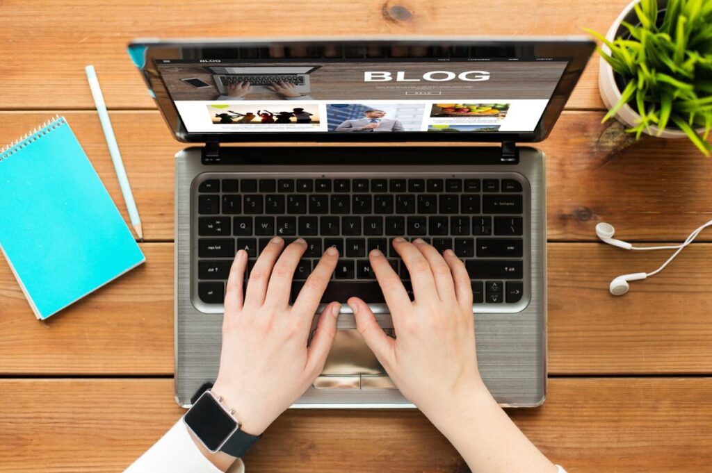 Close-up of a person typing on a laptop displaying a blog page, surrounded by a notebook, pencil, plant, and earphones on a wooden desk