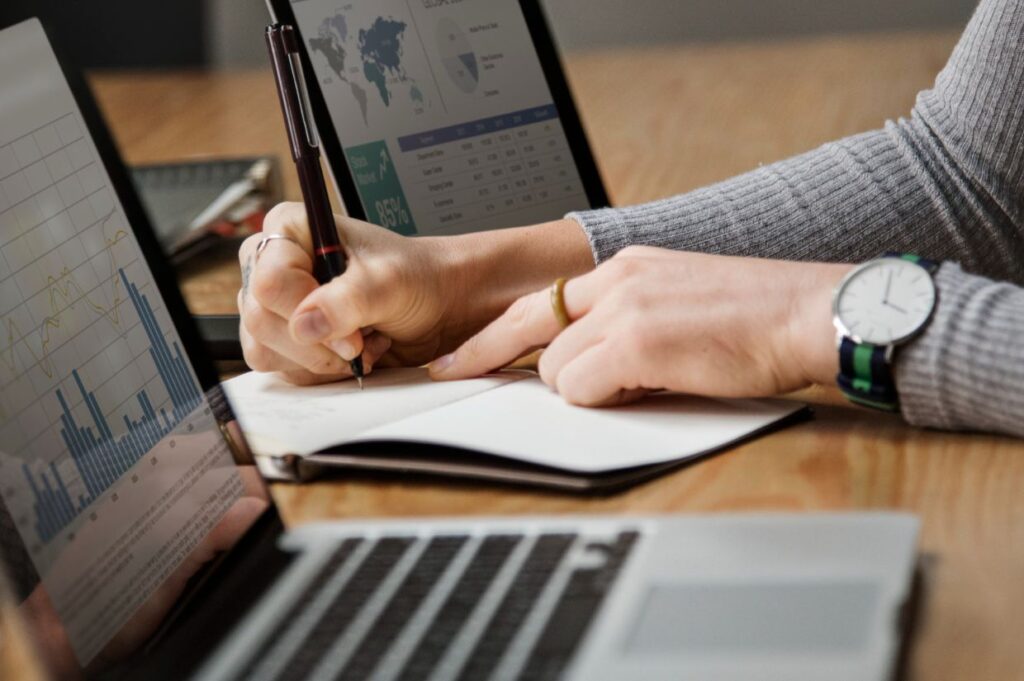 A person writing in a notebook while analyzing data on two laptops.