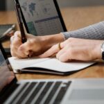 A person writing in a notebook while analyzing data on two laptops.