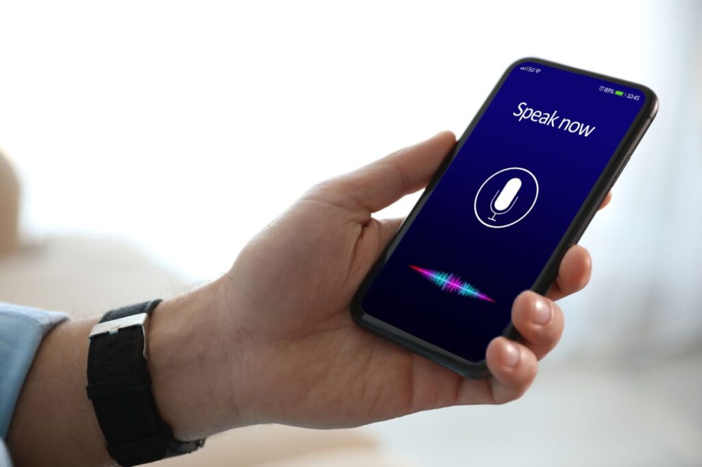 A close-up of a hand holding a smartphone displaying a voice assistant interface. The screen reads "Speak now" with a microphone icon and colorful sound waves below, indicating active voice input.