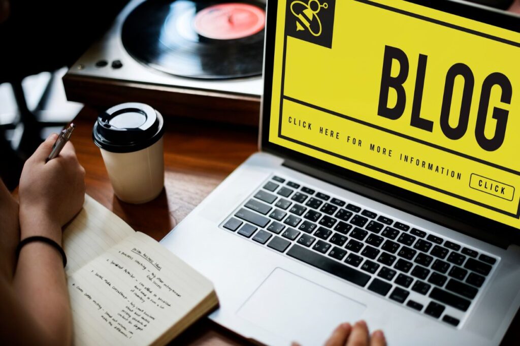 A person working on a blog, taking notes beside a laptop with a bold yellow "BLOG" screen, a coffee cup, and a vinyl player.