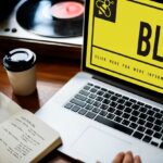 A person working on a blog, taking notes beside a laptop with a bold yellow "BLOG" screen, a coffee cup, and a vinyl player.