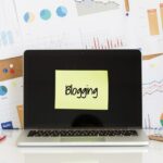 A laptop with a yellow sticky note on the screen that reads "Blogging" is positioned in front of a cluttered workspace.