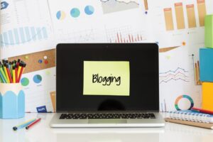 A laptop with a yellow sticky note on the screen that reads "Blogging" is positioned in front of a cluttered workspace.