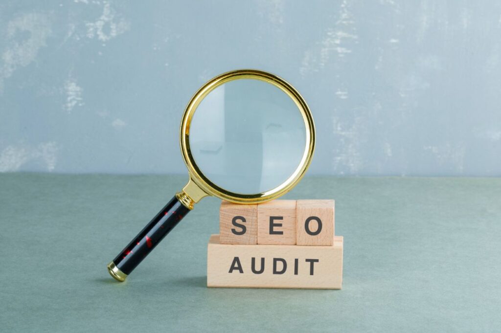 A magnifying glass is positioned over wooden blocks that spell out "SEO AUDIT," symbolizing the concept of SEO analysis or business evaluation.