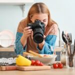 A woman with light brown hair is photographing a plated dish with a professional DSLR camera in a bright kitchen.