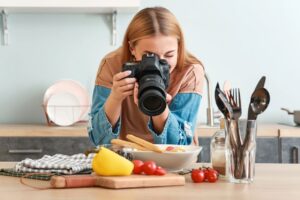 A woman with light brown hair is photographing a plated dish with a professional DSLR camera in a bright kitchen.