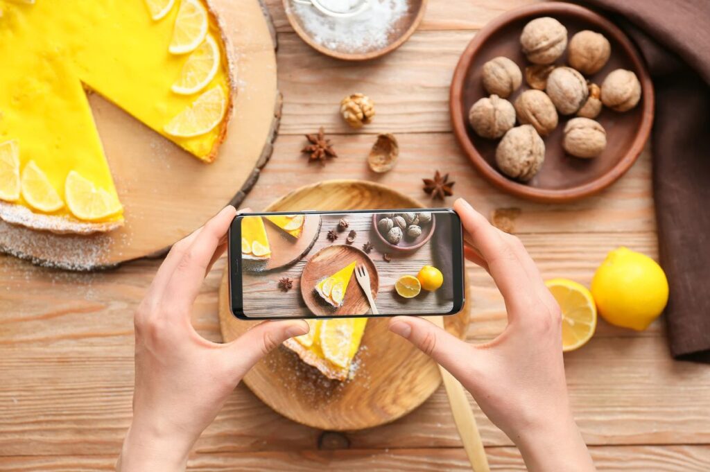 A person is holding a smartphone and taking a photo of a slice of lemon tart on a wooden plate. 