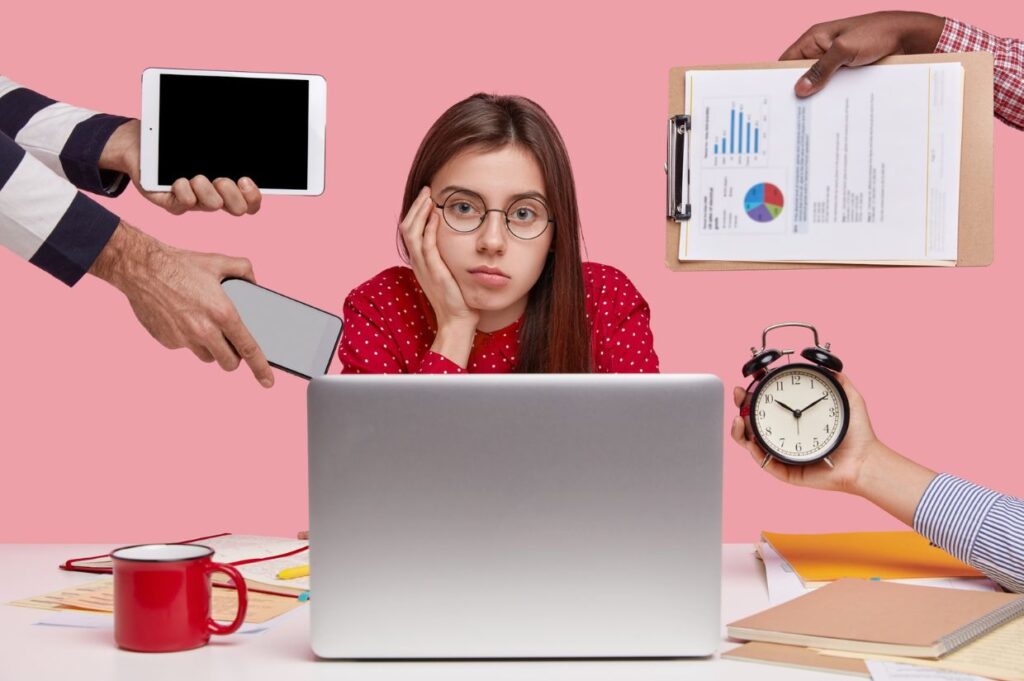 A young woman with long brown hair and glasses sits at a desk with a laptop, resting her face on her hand and looking overwhelmed.