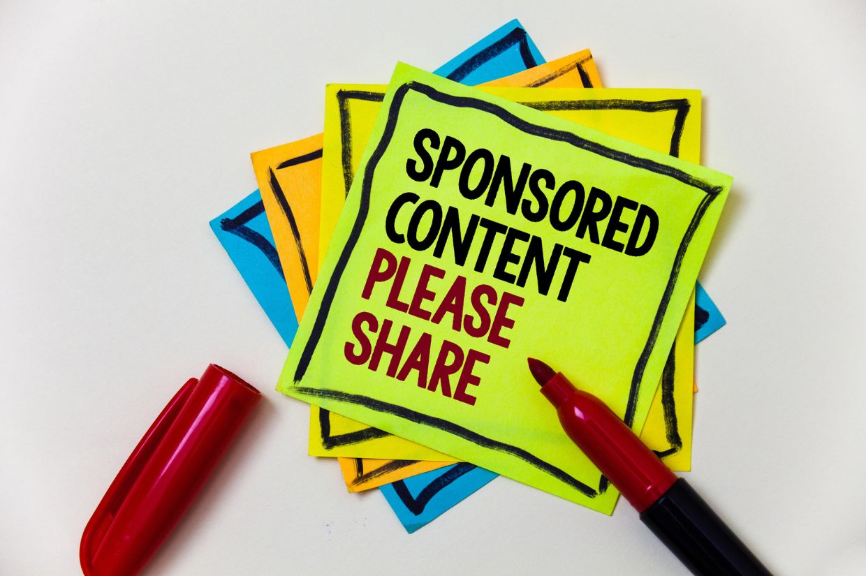 A stack of colorful sticky notes with the top note reading "SPONSORED CONTENT PLEASE SHARE" in bold letters, surrounded by a red marker. Represents the promotion and ethical considerations of sponsored content in media.