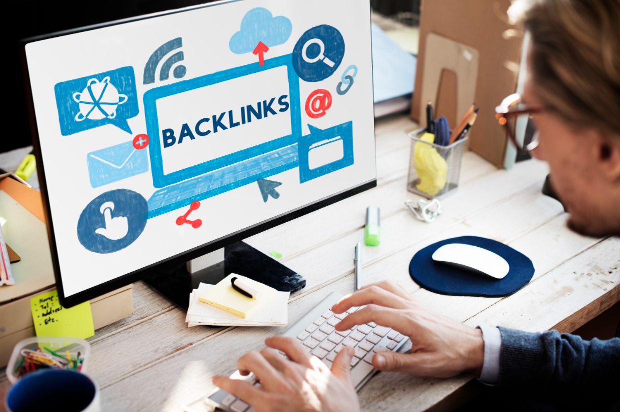 A person working at a desk on a computer with a screen displaying the word "BACKLINKS" surrounded by digital marketing icons, symbolizing strategies to build blog authority through backlinks in 2025.
